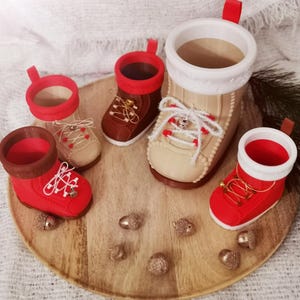 Könnte beinhalten: Eine Sammlung dekorativer stiefelförmiger Behälter in verschiedenen Größen und Farben. Die Stiefel sind rot, braun und beige, mit weißen Schnürsenkeln und roten Akzenten. Sie sind auf einem Holztablett mit goldfarbenen Eicheln angeordnet.