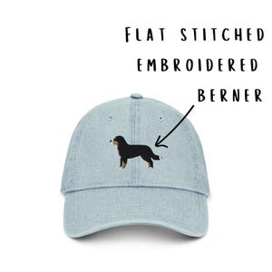 May include: A light blue denim baseball cap featuring a flat stitched embroidered Bernese Mountain Dog on the front. The text FLAT STITCHED EMBROIDERED BERNER is printed above the cap with an arrow pointing to the dog embroidery.