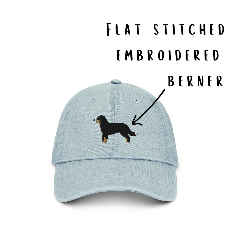 May include: A light blue denim baseball cap featuring a flat stitched embroidered Bernese Mountain Dog on the front. The text FLAT STITCHED EMBROIDERED BERNER is printed above the cap with an arrow pointing to the dog embroidery.