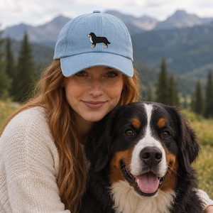 May include: A light blue baseball cap featuring an embroidered graphic of a black, white, and tan Bernese Mountain Dog on the front panel. The cap has a curved brim and a structured crown, showcasing a detailed canine illustration in a standing pose.