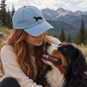 May include: A light blue baseball cap featuring an embroidered graphic of a black, white, and tan Bernese Mountain Dog on the front panel. The cap has a curved brim, a structured crown with eyelets, and a fabric strap closure at the back.