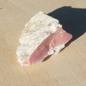 Op de afbeelding: Een ruwe roze en witte opaal edelsteen. De opaal heeft een glad, gepolijst oppervlak en een ruwe, natuurlijke achterkant.