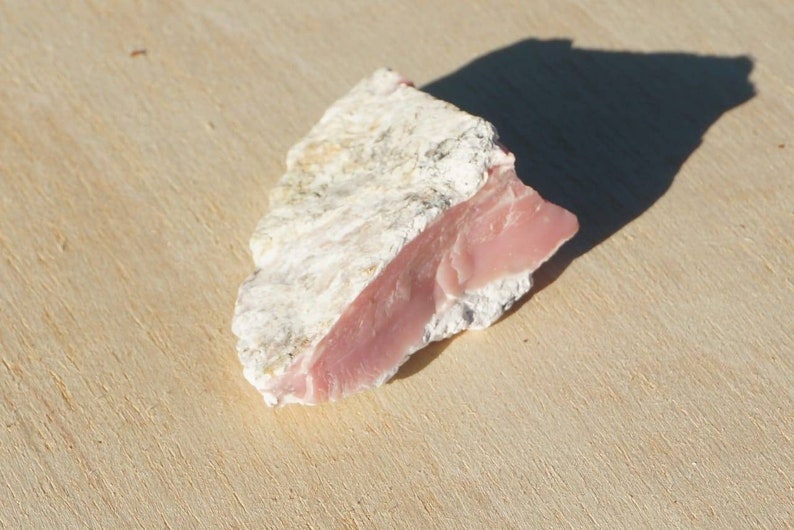 Op de afbeelding: Een ruwe roze en witte opaal edelsteen. De opaal heeft een glad, gepolijst oppervlak en een ruwe, natuurlijke achterkant.