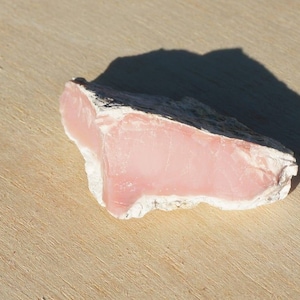 Op de afbeelding: Een ruwe, roze en witte, natuurlijke, ondoorzichtige, halfedelsteen. De steen heeft een ruwe, onregelmatige vorm en is lichtroze.