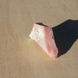 Pode incluir: Uma pe&ccedil;a de opala rosa bruta de corte rosa e branco. A opala tem uma forma triangular e uma textura &aacute;spera e natural.