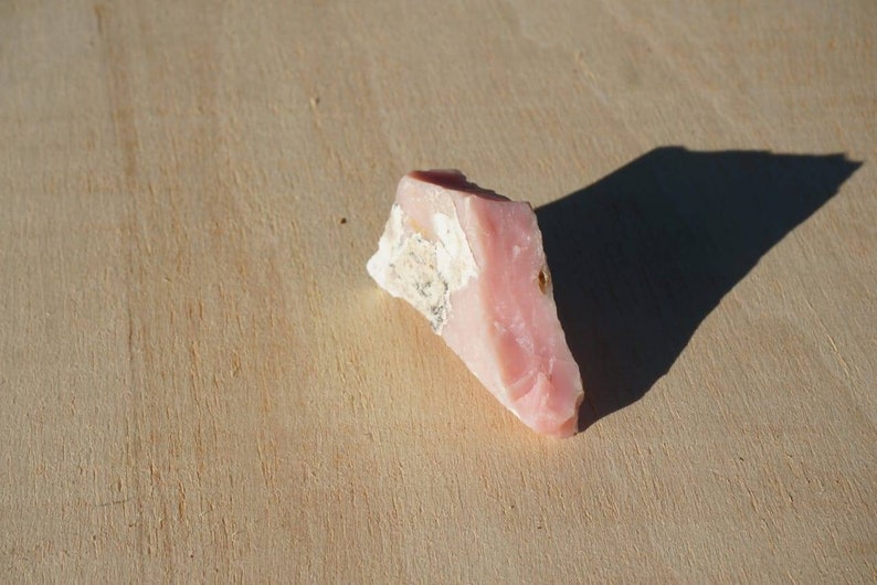 Pode incluir: Uma pe&ccedil;a de opala rosa bruta de corte rosa e branco. A opala tem uma forma triangular e uma textura &aacute;spera e natural.