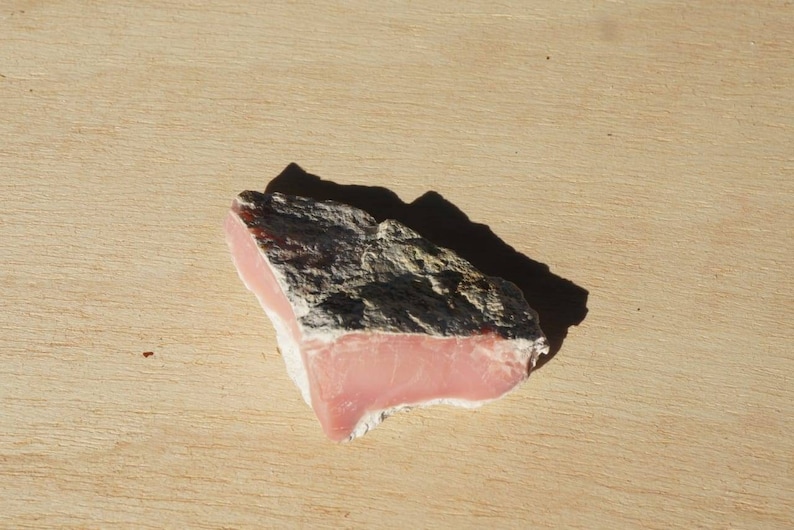 Op de afbeelding: Een ruw, grijs en roze, onregelmatig gevormd stuk roze opaal. De opaal ligt op een lichtbruin houten oppervlak.