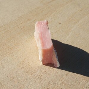 Pode incluir: Um cristal de opala rosa com uma textura &aacute;spera e natural. O cristal &eacute; de cor rosa claro e tem uma apar&ecirc;ncia transl&uacute;cida.