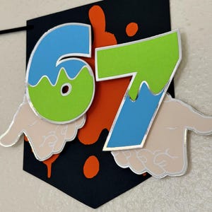 May include: A black banner with the numbers "67" in blue and green, outlined in silver. The banner has orange paint splatters and stylized hands on the bottom. The banner is suspended by a black string.