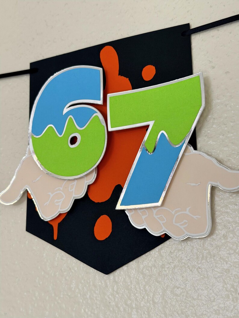 May include: A black banner with the numbers "67" in blue and green, outlined in silver. The banner has orange paint splatters and stylized hands on the bottom. The banner is suspended by a black string.