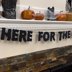 Puede incluir: Una pancarta de Halloween con el texto "HERE FOR THE BOOS" en letras negras que gotean. La pancarta está adornada con recortes temáticos de Halloween, incluyendo un fantasma, un cóctel y copas de vino. La pancarta se muestra sobre una chimenea decorada con calabazas y un esqueleto.