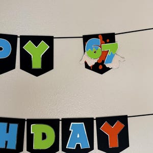 May include: A black banner with the words "HAPPY BIRTHDAY" in blue, green, and orange letters. The number "67" is displayed on a separate banner with a splatter design and hand-shaped cutouts.