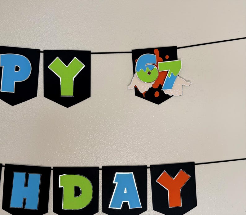 May include: A black banner with the words "HAPPY BIRTHDAY" in blue, green, and orange letters. The number "67" is displayed on a separate banner with a splatter design and hand-shaped cutouts.