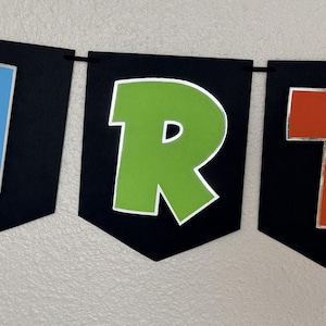 May include: A banner with black pennants displaying colorful letters. The letter "R" is green with a silver outline. The banner is strung on a black string and is against a white wall.
