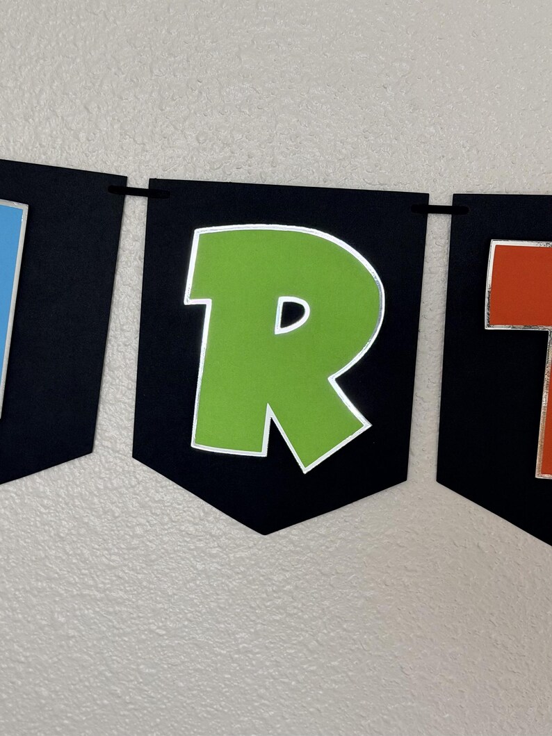 May include: A banner with black pennants displaying colorful letters. The letter "R" is green with a silver outline. The banner is strung on a black string and is against a white wall.