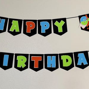 May include: A black banner with the words "HAPPY BIRTHDAY" in colorful letters. The letters are outlined in white and have a 3D effect. A separate banner displays the number "67". The banner is strung on a black string.