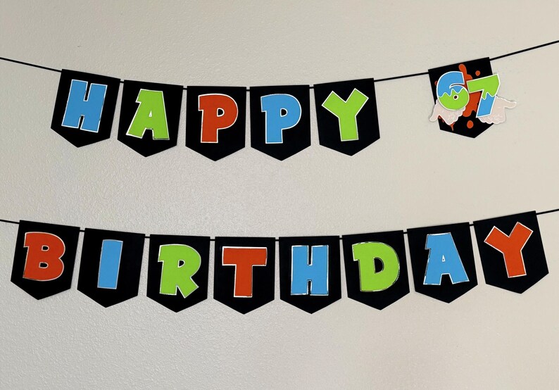 May include: A black banner with the words "HAPPY BIRTHDAY" in colorful letters. The letters are outlined in white and have a 3D effect. A separate banner displays the number "67". The banner is strung on a black string.