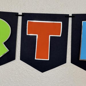 May include: A banner with black pennants displaying colorful letters. The letter "T" is orange with a silver outline, set against a black background. The banner is strung with a black cord, suitable for party decorations.