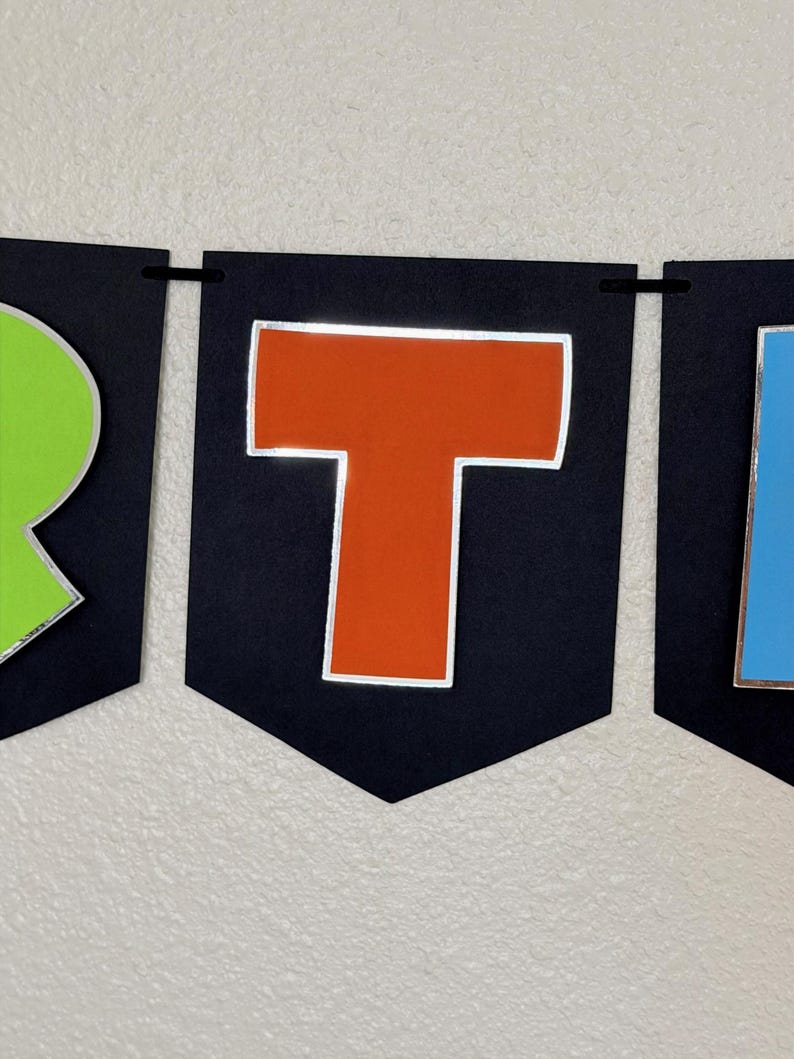 May include: A banner with black pennants displaying colorful letters. The letter "T" is orange with a silver outline, set against a black background. The banner is strung with a black cord, suitable for party decorations.