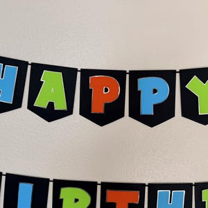 May include: A black banner with the words "HAPPY BIRTHDAY" in colorful letters. The letters are outlined in silver and have a 3D effect. The banner is strung on a black string, perfect for a birthday party.