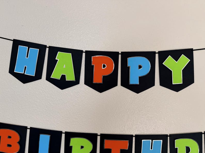 May include: A black banner with the words "HAPPY BIRTHDAY" in colorful letters. The letters are outlined in silver and have a 3D effect. The banner is strung on a black string, perfect for a birthday party.
