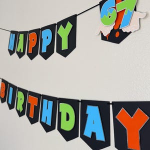 May include: A black banner with the words "HAPPY BIRTHDAY" in colorful letters. The letters are outlined in silver and have a 3D effect. A separate banner displays the number "67" with a splatter design.