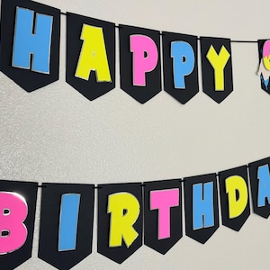 May include: A black banner with the words "HAPPY BIRTHDAY" in colorful, 3D letters. The letters are outlined in silver and have a blue, pink, and yellow color scheme. A small banner with the number "67" is also included.