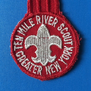 Peut inclure: Écusson brodé rouge avec une fleur de lys blanche au centre. Le texte autour de la fleur de lys se lit comme suit : "Twin Valley River Scout Greater New York".