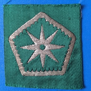 May include: A green fabric patch with a silver embroidered starburst design. The starburst is in the shape of a pentagon with a star in the centre.