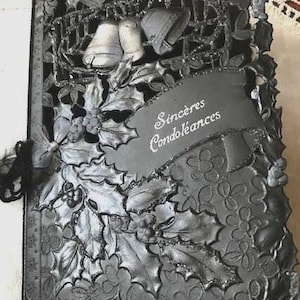 May include: A grey, embossed sympathy card with a floral and bell design. The card features the words "Sincères Condoléances" on a banner. The card is decorated with holly leaves and berries, and a black ribbon.