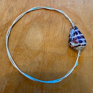 Peut inclure: Un bracelet fait à la main avec un fil d'argent et un pendentif en forme de goutte d'eau. Le coquillage est marron, blanc et bleu, ajoutant un élément naturel. Le bracelet est conçu pour être porté autour du poignet.
