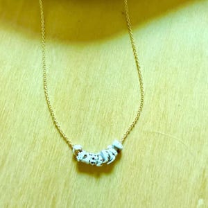 May include: A delicate gold-tone necklace featuring a series of white and black speckled beads. The beads are strung together in a curved line, suspended from a fine chain. The necklace is displayed against a light yellow background.