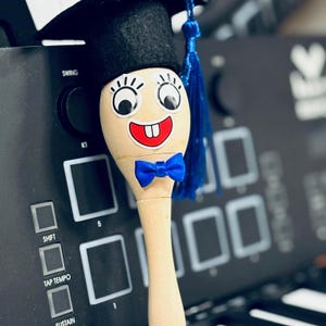 Pode incluir: Uma colher de pau com um rosto de desenho animado, uma gravata borboleta azul e um pequeno chapéu de formatura. A colher está posicionada em frente a um dispositivo eletrônico preto com botões e um mostrador.