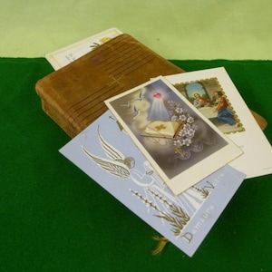 May include: A collection of religious items including a brown leather-bound book with a cross, and several vintage postcards. The postcards feature religious imagery, including angels and biblical scenes. The items are arranged on a green surface.