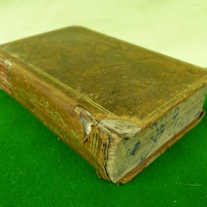 May include: A well-worn, rectangular book with a brown leather cover. The edges show signs of age and wear, with gold detailing and visible text. The book is resting on a green surface.