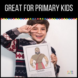 May include: A young boy holds up a printable educational card featuring a cartoon illustration of Goliath from the Bible. The card has the text "Goliath" and a quote about Goliath's height and challenge to the Israelites. The card is part of a set for primary school children.