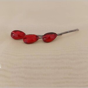 Puede incluir: Tres horquillas rojas en forma de l&aacute;grima con tallos plateados se muestran sobre una superficie beige claro. Las horquillas est&aacute;n dispuestas en una l&iacute;nea ligeramente escalonada, mostrando su color vibrante y su delicado dise&ntilde;o. El logotipo de DENMARBG est&aacute; en la esquina inferior izquierda.