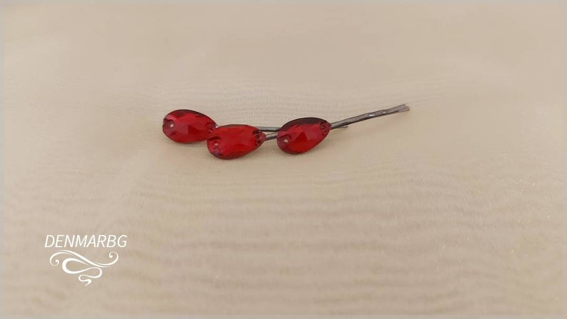 Puede incluir: Tres horquillas rojas en forma de l&aacute;grima con tallos plateados se muestran sobre una superficie beige claro. Las horquillas est&aacute;n dispuestas en una l&iacute;nea ligeramente escalonada, mostrando su color vibrante y su delicado dise&ntilde;o. El logotipo de DENMARBG est&aacute; en la esquina inferior izquierda.