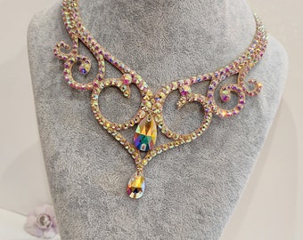Puede incluir: Un collar de tono dorado con un diseño delicado y ornamentado que presenta un colgante de cristal en forma de lágrima. El collar está adornado con diamantes de imitación iridiscentes.
