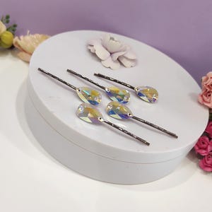 Puede incluir: Seis horquillas para el pelo con cristales iridiscentes en forma de l&aacute;grima. Las horquillas tienen tallos de metal oscuro y est&aacute;n dispuestas sobre una superficie redonda blanca. Un fondo lila claro y acentos florales completan la imagen.