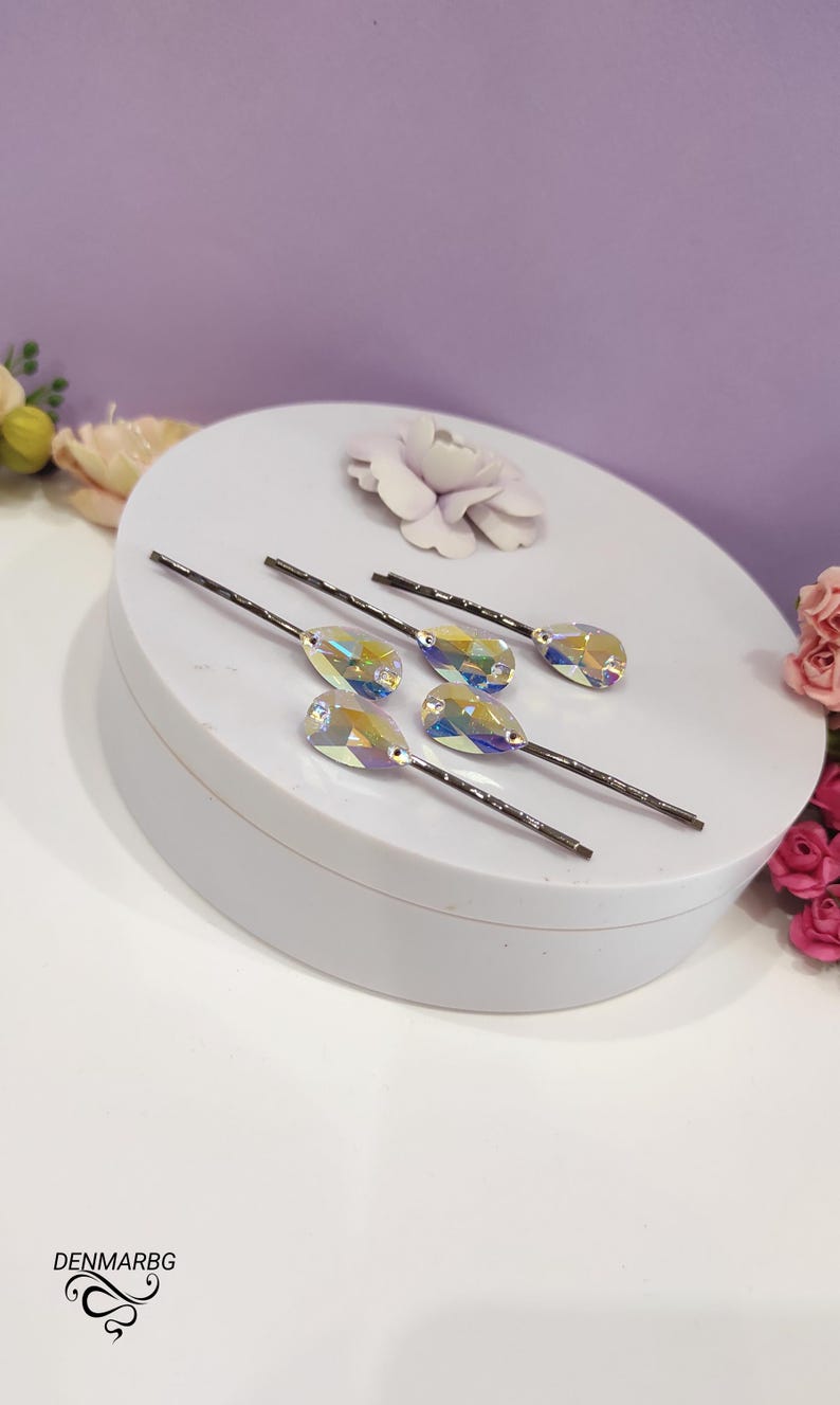 Puede incluir: Seis horquillas para el pelo con cristales iridiscentes en forma de l&aacute;grima. Las horquillas tienen tallos de metal oscuro y est&aacute;n dispuestas sobre una superficie redonda blanca. Un fondo lila claro y acentos florales completan la imagen.