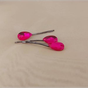 Puede incluir: Tres horquillas rosas fucsia con joyas en forma de l&aacute;grima. Las horquillas son de color plateado y las joyas son de un color fucsia vibrante. Las horquillas est&aacute;n dispuestas sobre una superficie de tono neutro.