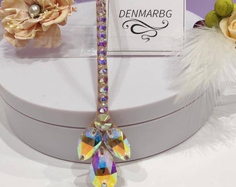 May include: A crystal headband with a row of iridescent rhinestones and a cluster of three larger iridescent crystals at the end. The headband is on a white surface with a white feather and a flower.