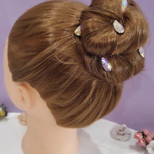 Puede incluir: Una cabeza de maniqu&iacute; con cabello casta&ntilde;o peinado en un mo&ntilde;o, adornada con varias horquillas de cristal iridiscente. El cabello est&aacute; peinado con esmero y las horquillas a&ntilde;aden un toque de brillo. El fondo es de color p&uacute;rpura suave.