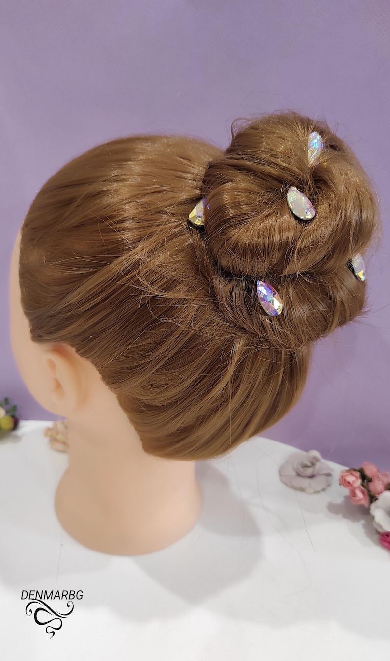Puede incluir: Una cabeza de maniqu&iacute; con cabello casta&ntilde;o peinado en un mo&ntilde;o, adornada con varias horquillas de cristal iridiscente. El cabello est&aacute; peinado con esmero y las horquillas a&ntilde;aden un toque de brillo. El fondo es de color p&uacute;rpura suave.