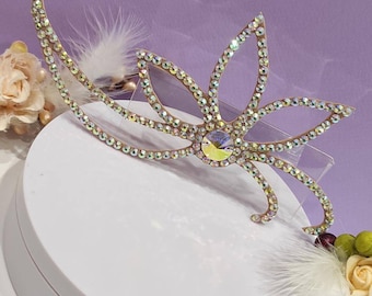 May include: A gold-toned, crystal-encrusted hairpiece with a flower design. The hairpiece is on a white circular surface with white feathers.