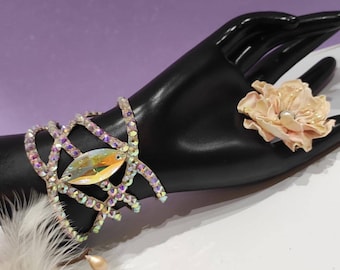 May include: A crystal bracelet with a large iridescent stone in the center. The bracelet is on a black mannequin hand with a pink flower on the finger.