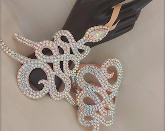 May include: A pair of ornate, crystal-encrusted, snake-shaped arm cuffs. The cuffs are made of a light brown material and feature a delicate, intricate design. The cuffs are adorned with iridescent crystals that sparkle in the light.