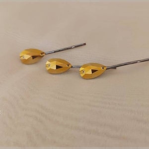 Puede incluir: Tres horquillas doradas con detalles de cristal facetado en forma de l&aacute;grima. Las horquillas tienen una base de metal plateado. El fondo es de color beige claro. La marca "DENMARBG" est&aacute; en la esquina inferior izquierda.