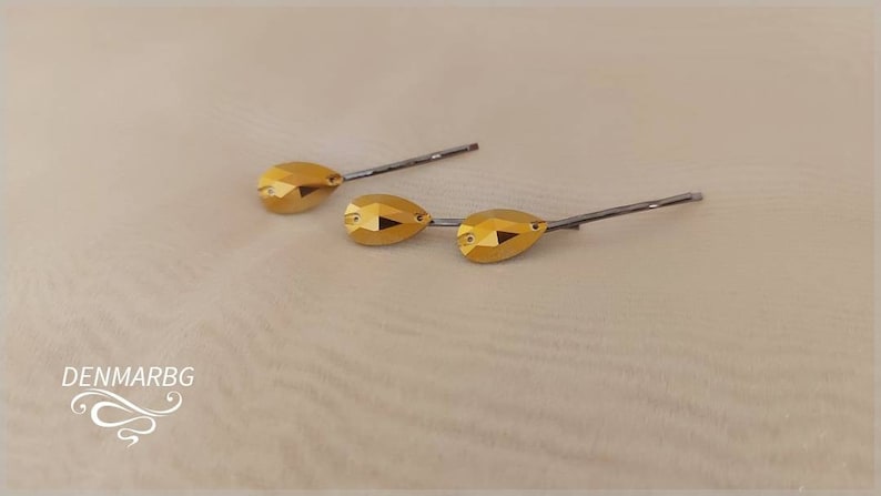 Puede incluir: Tres horquillas doradas con detalles de cristal facetado en forma de l&aacute;grima. Las horquillas tienen una base de metal plateado. El fondo es de color beige claro. La marca "DENMARBG" est&aacute; en la esquina inferior izquierda.